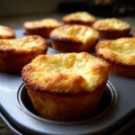 Close-up of golden-brown baby lemon impossible pies baked in a muffin tin, showing their slightly puffed tops.