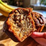 Close-up of a moist banana nut muffin, broken in half to show the texture and visible walnuts. Another muffin is in the background.