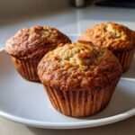 Three golden-brown banana protein muffins displayed on a white plate, ready to eat.