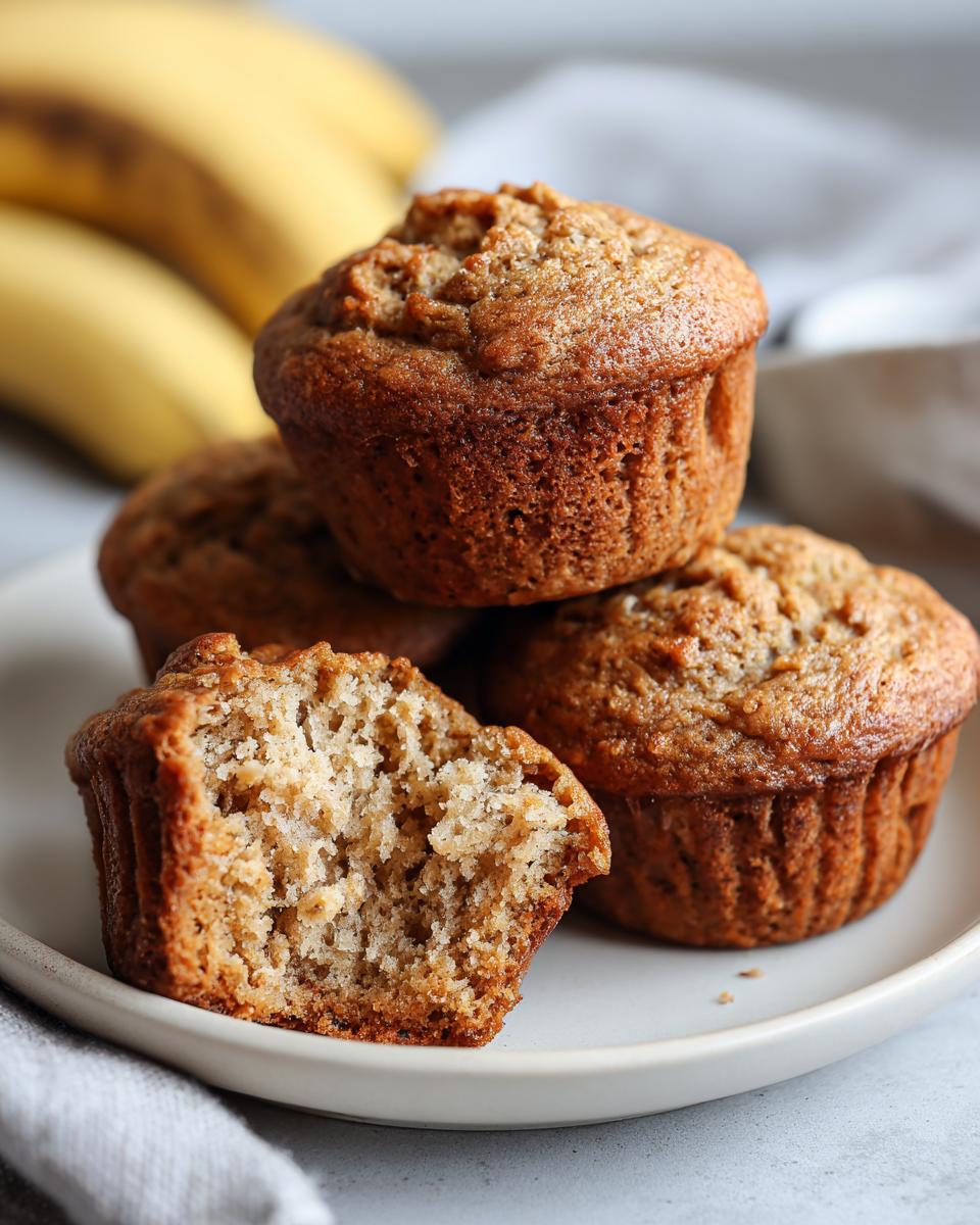 A stack of golden brown banana protein muffins on a white plate, with one muffin broken in half to show the fluffy interior.