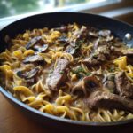 Close-up of a pan filled with creamy Beef Stroganoff, featuring tender beef strips, mushrooms, and egg noodles, garnished with herbs.