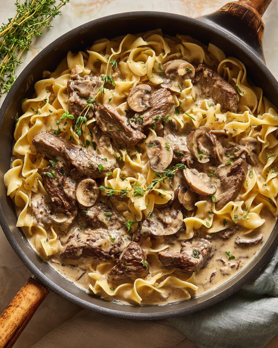 Close-up of a skillet filled with creamy beef stroganoff and egg noodles, garnished with fresh thyme.