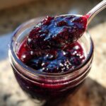 A spoonful of glistening homemade blueberry jam being lifted from a glass jar.