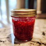 Close-up of a glass jar filled with vibrant red Christmas jam, showing fruit pieces and seeds.