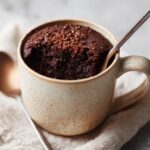 A close-up of a rich chocolate Cottage Cheese Microwave Brownie in a speckled mug, with a spoon taking a bite.