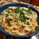 A close-up of a bowl of creamy lemon pasta with shredded chicken and fresh parsley.