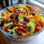 A close-up of a large glass bowl filled with a colorful Dorito Taco Salad, featuring seasoned ground beef, lettuce, tomatoes, olives, and crushed Doritos.