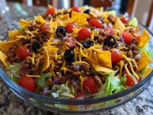 A close-up of a large glass bowl filled with Dorito Taco Salad, featuring lettuce, seasoned ground beef, shredded cheese, cherry tomatoes, black olives, and crushed Doritos.