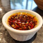 Close-up of a small white bowl filled with flavorful dumpling sauce, featuring chili flakes and sesame seeds.