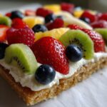 Close-up of a slice of fruit pizza on a sugar cookie crust, topped with cream cheese and fresh berries and kiwi.