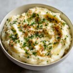 A close-up of a bowl of creamy garlic parmesan mashed potatoes, topped with melted butter, parsley, and paprika.