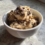 A close-up of a white bowl filled with creamy, high protein cookie dough studded with dark chocolate chips.