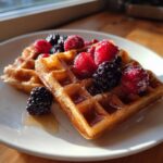 Two golden-brown high protein waffles topped with fresh raspberries and blackberries, drizzled with syrup.
