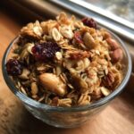 A close-up of a glass bowl filled with delicious homemade granola, featuring oats, nuts, seeds, and dried cranberries.