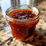 Close-up of a glass jar filled with glossy, rich honey BBQ sauce, showing its thick texture and flecks of spices.