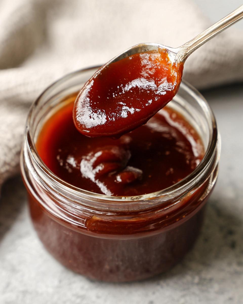 A spoonful of rich, glossy honey BBQ sauce being lifted from a glass jar.