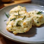 Three fluffy rosemary biscuits topped with fresh rosemary sprigs, served on a grey plate. Perfect with garlic aioli.