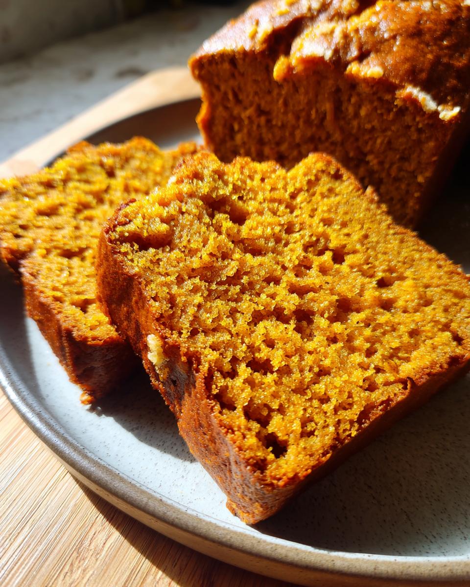 Close-up of two moist slices of sourdough pumpkin bread on a plate, with the loaf in the background.
