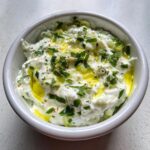 Close-up of creamy homemade Tzatziki sauce in a white bowl, drizzled with olive oil and sprinkled with fresh herbs.