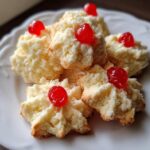 A close-up of melt-in-mouth whipped shortbread cookies, topped with bright red cherries, on a white plate.