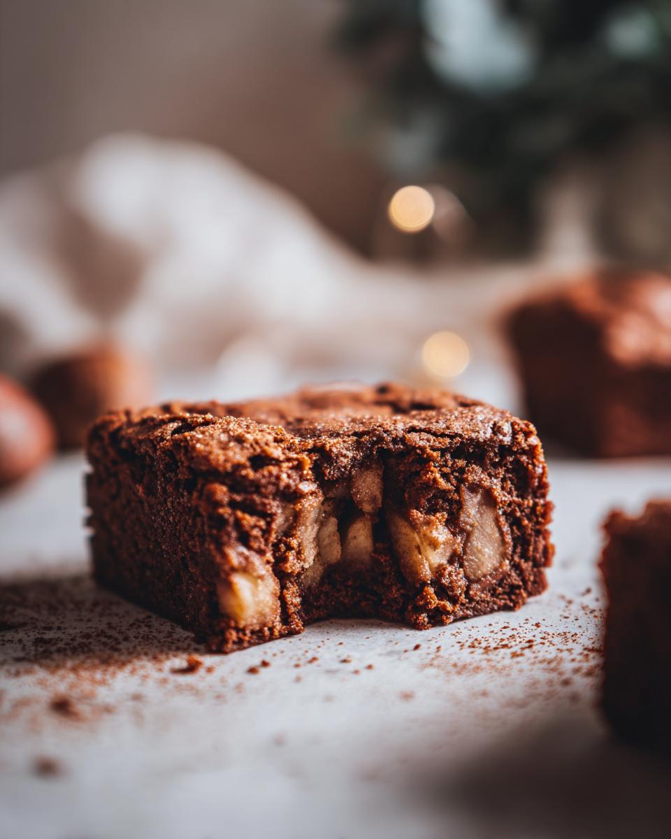 A close-up of a fudgy apple brownie with chunks of apple visible, dusted with cocoa powder.