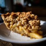 A close-up of a slice of apple crisp pie on a white plate, showing tender apple filling and a crumbly topping.