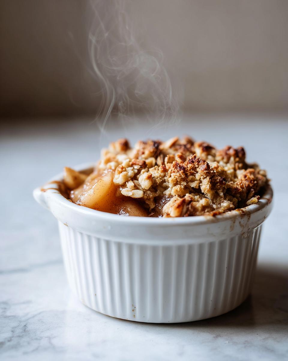 A close-up shot of a steaming individual apple crisp in a white ramekin, with a golden-brown crumble topping.