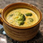 A close-up of creamy Broccoli Cheddar Soup served in a rustic brown mug, garnished with fresh broccoli florets and melted cheddar cheese.