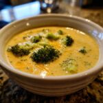 A close-up of a white bowl filled with creamy broccoli potato cheese soup, topped with fresh broccoli florets and shredded cheese.