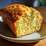 A close-up of a golden-brown Cheddar Herb Quick Bread loaf on a plate, showing visible chunks of cheddar and herbs.