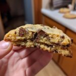 Close-up of a hand holding a perfectly chewy chocolate chip cookie, showing its soft interior and melted chocolate chunks.