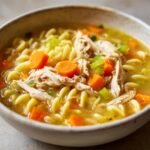 A close-up shot of a comforting bowl of chicken noodle soup, filled with shredded chicken, carrots, celery, and pasta.