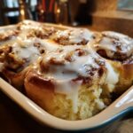 Close-up of fresh baked cinnamon rolls generously topped with cream cheese glaze in a baking dish.