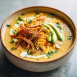 A close-up of a bowl of creamy chicken tortilla soup topped with shredded cheese, crispy tortilla strips, avocado, and cilantro.