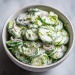 A bowl of creamy cucumber salad featuring thinly sliced cucumbers, red onion rings, and fresh dill.
