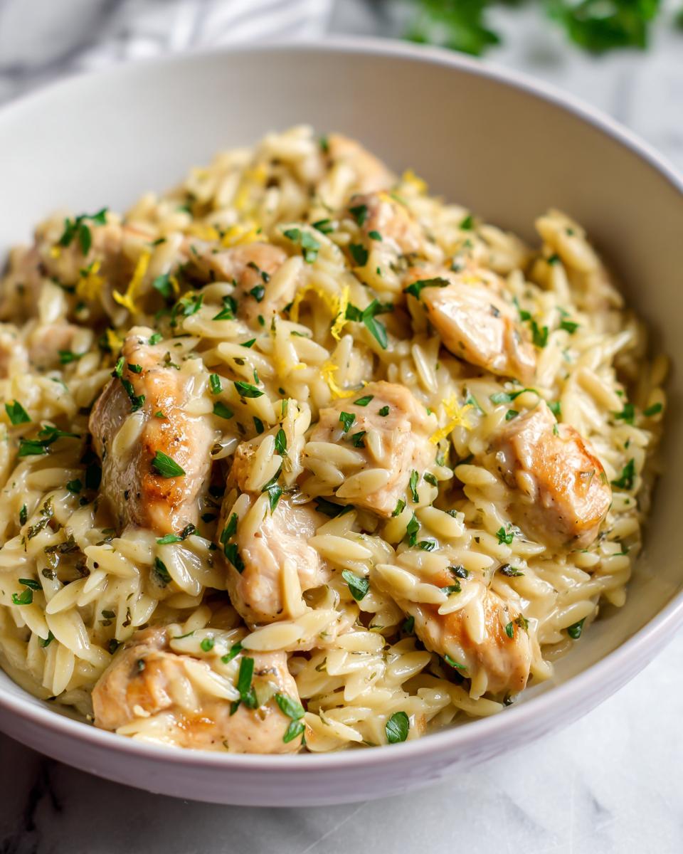 A close-up of creamy lemon chicken orzo, featuring tender chicken pieces and orzo pasta in a rich sauce, garnished with fresh parsley and lemon zest.