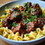 A close-up of a white bowl filled with tender crockpot beef tips and noodles, garnished with fresh parsley.