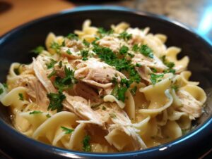 A close-up of a dark bowl filled with creamy crockpot chicken and noodles, garnished with fresh parsley.