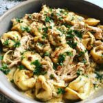 Close-up of a bowl filled with creamy Crockpot Chicken Tortellini, garnished with fresh parsley and red pepper flakes.