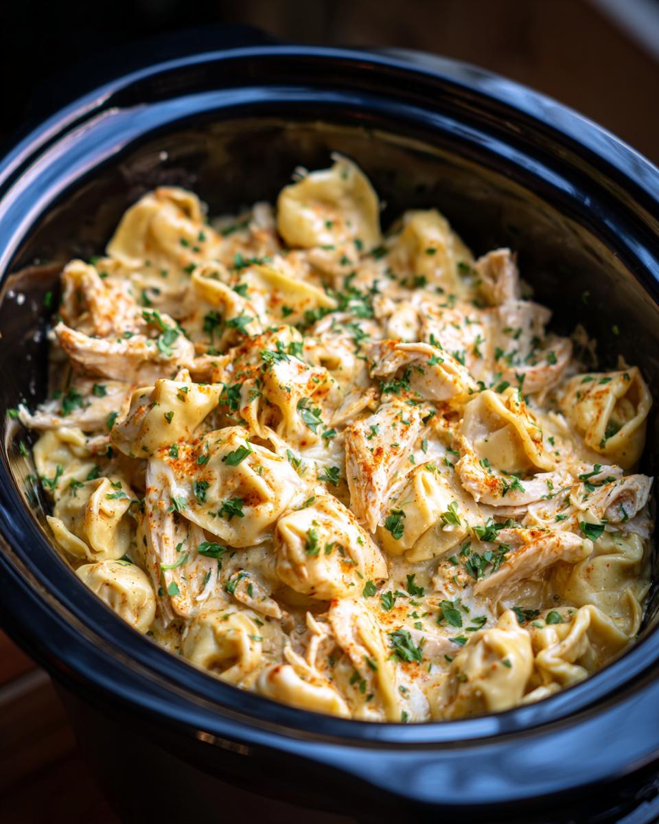 Close-up of creamy Crockpot Chicken Tortellini garnished with parsley and paprika in a black slow cooker.