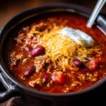 A close-up of a bowl of hearty Crockpot chili topped with shredded cheese and a dollop of sour cream.