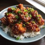A plate of crispy General Tso's chicken pieces served over white rice, garnished with sesame seeds and green onions.