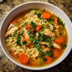 A steaming bowl of Ginger Garlic Chicken Noodle Soup with tender chicken, ramen noodles, carrots, celery, and parsley.