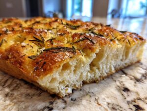 Close-up of a thick slice of golden-brown gluten free focaccia bread topped with fresh rosemary and coarse sea salt.