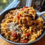A close-up shot of a fork lifting a portion of ground beef orzo with tomatoes and melted cheese.