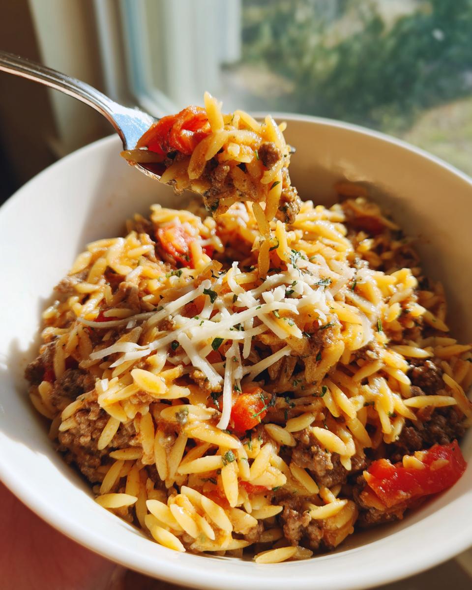 A fork lifts a bite of delicious ground beef orzo pasta with diced tomatoes and shredded cheese.
