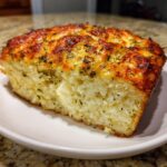 A close-up of a golden-brown slice of Italian herb and cheese bread, showing a fluffy interior and melted cheese topping.