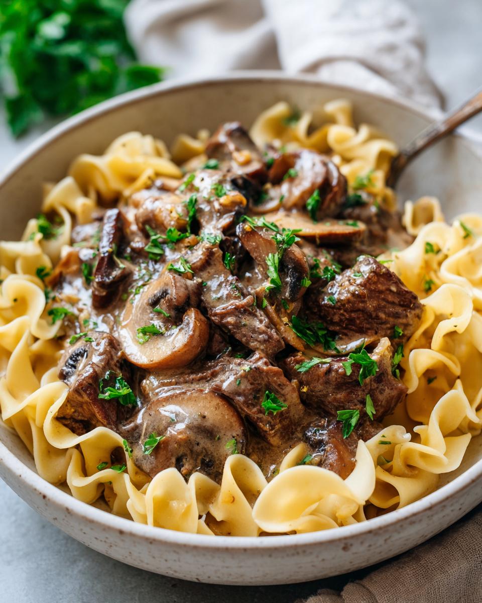 A close-up of a bowl filled with old fashioned beef stroganoff served over egg noodles, topped with fresh parsley.