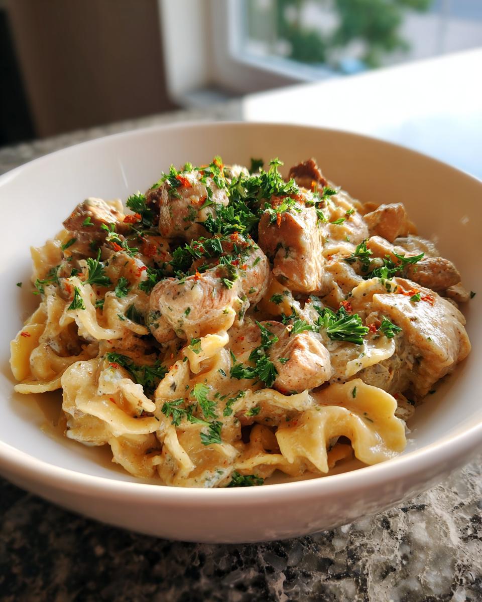 A close-up of a white bowl filled with creamy one-pan chicken and noodles, garnished with fresh parsley and red pepper flakes.