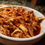 A close-up of shredded slow cooker bbq chicken coated in rich, glossy bbq sauce in a white bowl.